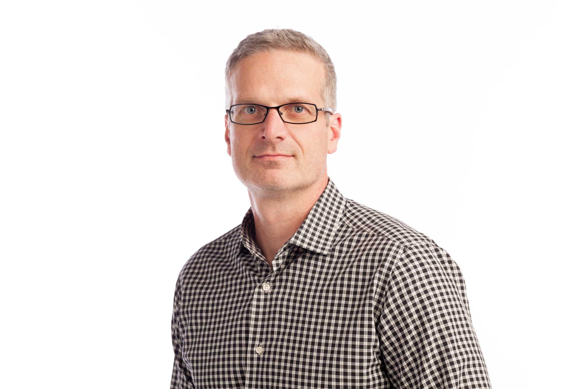 Studio headshot of Bryant King on a white background — short grey hair, black-framed glasses, black-and-white gingham button-down.
